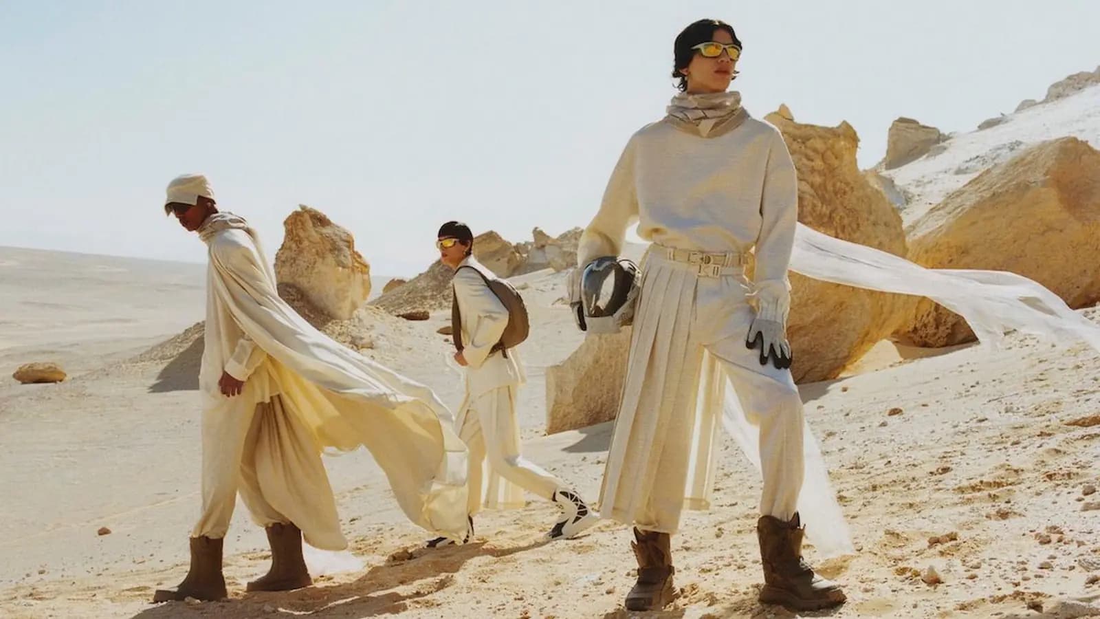 Dior Men Magazine Fall 2023 poster showing three fashion models in white desert styling walking through a rocky landscape.