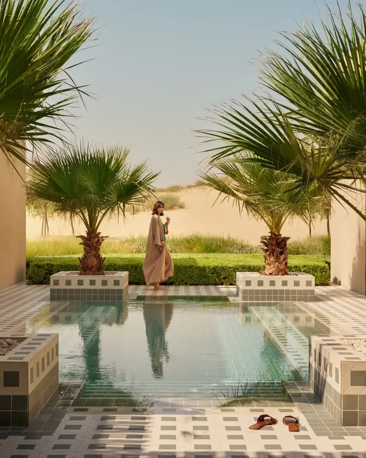 Bab Al Shams poster showing a woman walking beside a reflective pool framed by palm trees and desert landscaping.