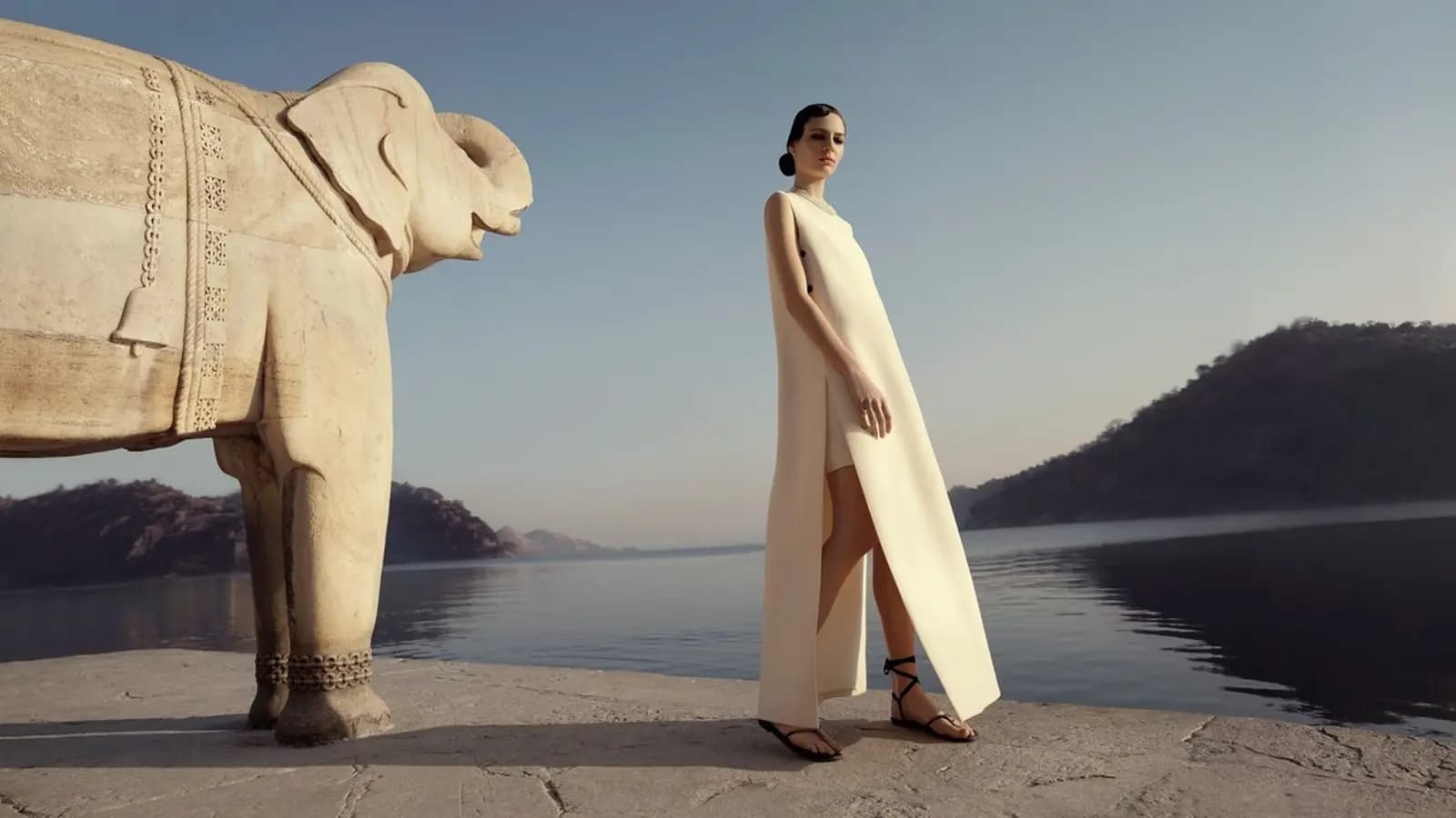 Dior Fall 2023 Women's Campaign poster showing a fashion model in a white dress standing beside a carved elephant sculpture near water and hills.