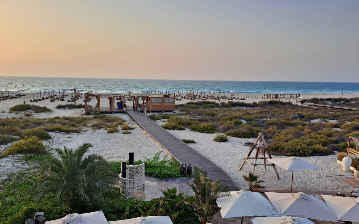 Abu Dhabi island beach setting with boardwalk, white sand, dunes, lounge areas, and Gulf water at sunset.