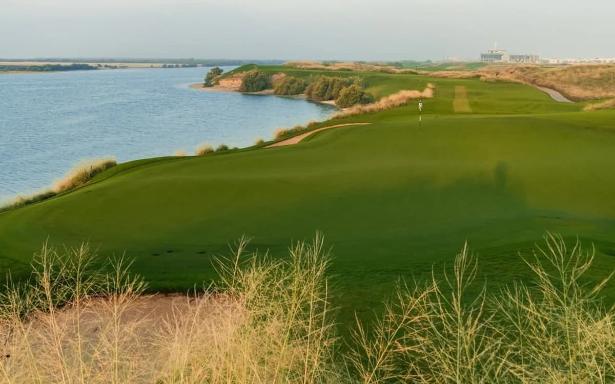 Abu Dhabi waterfront golf landscape with green fairway, water, coastline, and distant city or resort setting.