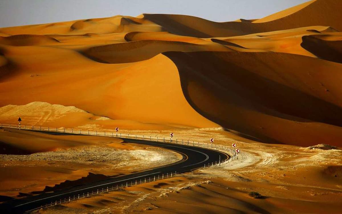 Desert road curving through Abu Dhabi dunes with high orange sand formations, road signs, and open inland terrain.