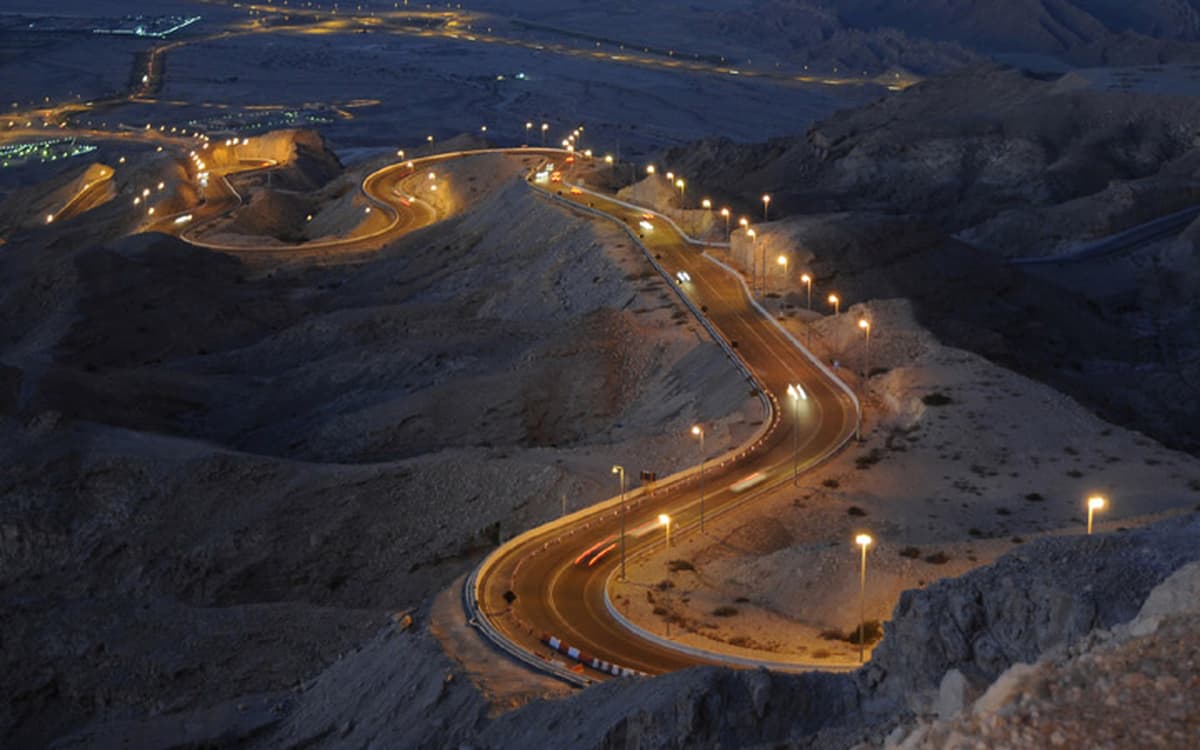 Mountain road in Abu Dhabi inland terrain at dusk with lit switchbacks, rocky slopes, and distant city lights.