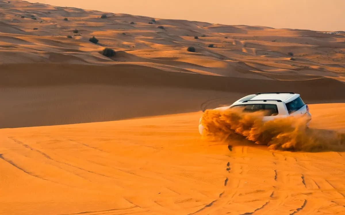 Off-road vehicle driving through Dubai desert dunes with orange sand, dust trail, and open desert landscape.