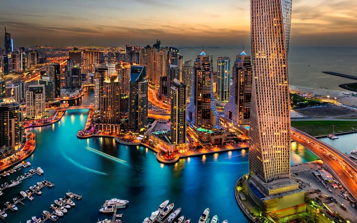 Dubai Marina skyline at sunset with high-rise towers, waterfront canals, yachts, roads, and coastal city lights.