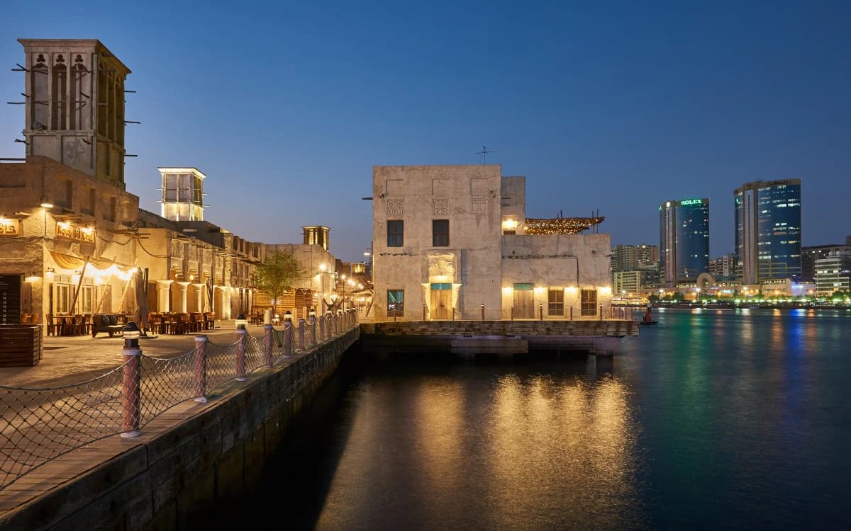 Dubai Creek at night with traditional wind-tower buildings, waterfront walkways, water reflections, and Deira skyline.