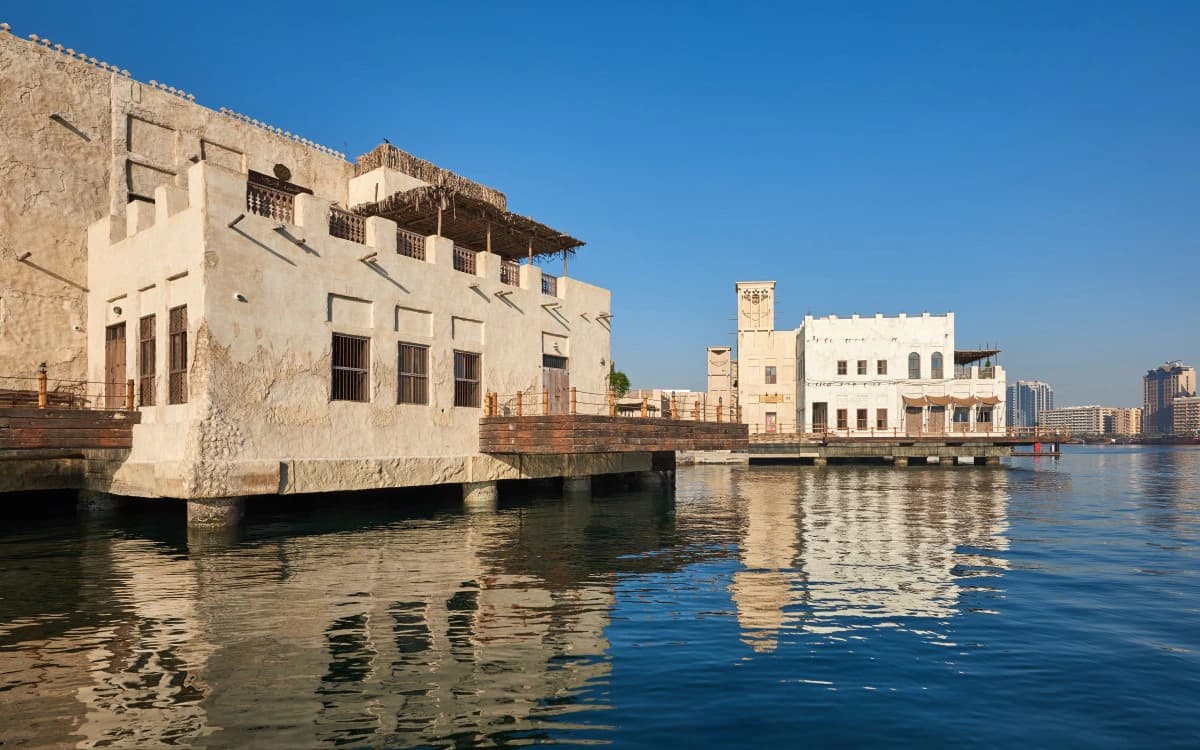 Old Dubai waterfront along Dubai Creek with traditional-style buildings, wind towers, wooden terraces, and calm water.