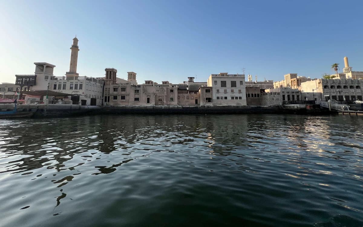 View across Dubai Creek toward old waterfront buildings, mosque minarets, wind towers, and water reflections.