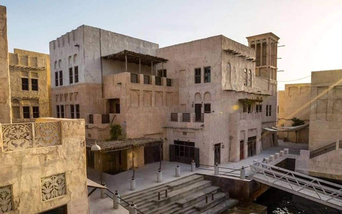 Old Dubai heritage-style waterfront buildings with wind towers, bridge, steps, and creek water at golden hour.