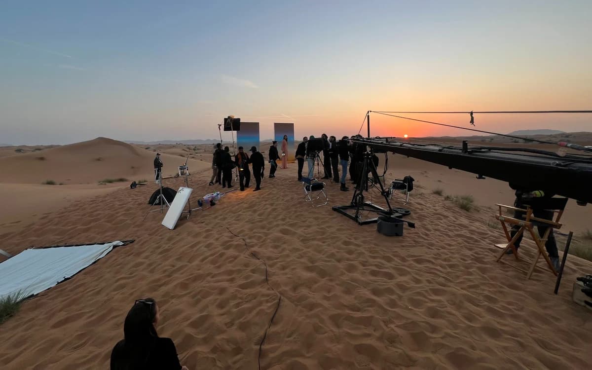 Desert film production setup in the UAE with crew, camera crane, lighting equipment, reflectors, and dunes at sunset.