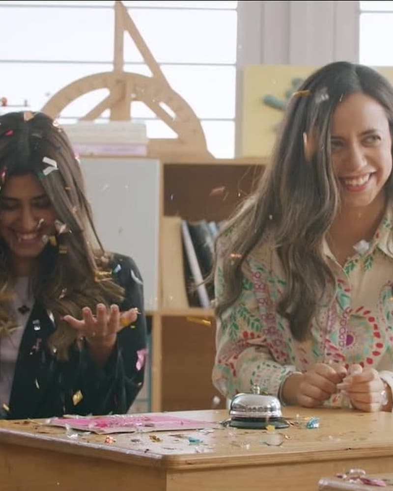 Two featured talent seated at classroom desks in an Amman launch episode set with confetti, bookshelves, and pastel classroom styling.