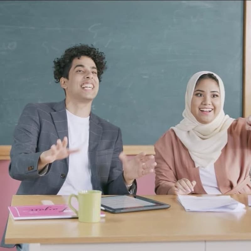 Two featured talent seated at a classroom desk in an Amman launch episode set with chalkboard, notebooks, and pastel school styling.