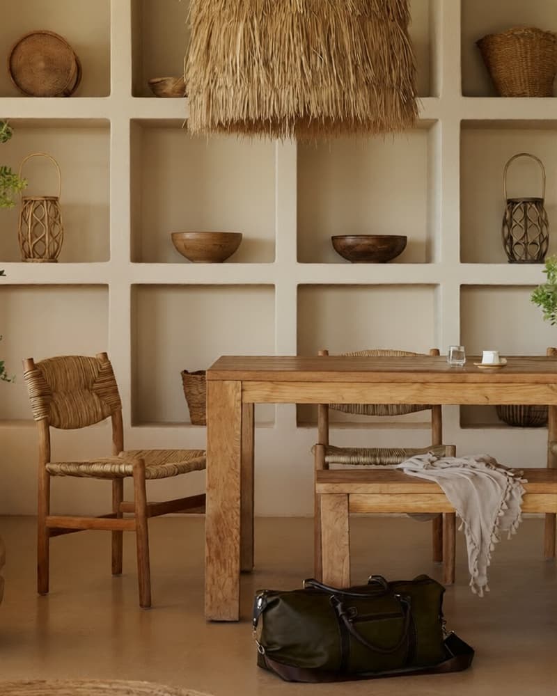 Styled hotel dining interior in Dubai with wooden table, woven chairs, shelving, baskets, pendant lamp, and travel bag.