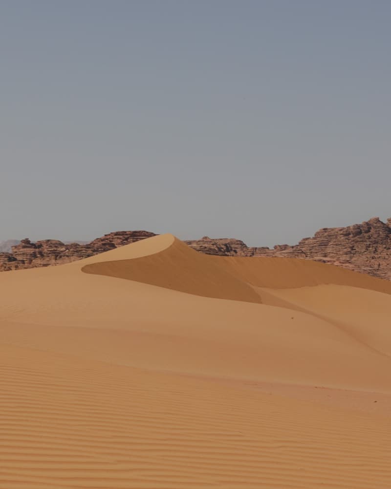 Wide desert landscape with vehicle tracks, open sand, distant mountains, and scattered clouds over remote terrain.