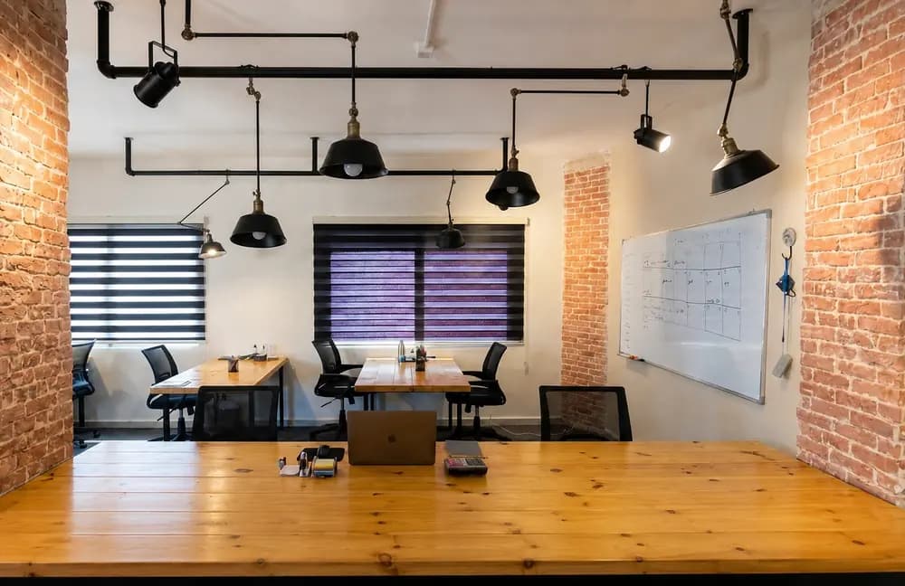 Versana Productions Amman office interior with wooden desks, exposed brick columns, black pendant lights, chairs, laptop, and whiteboard.