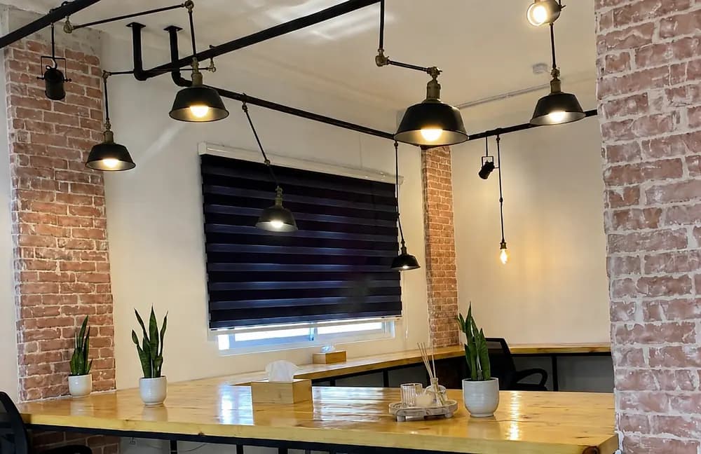 Amman office workspace with long wooden table, exposed brick columns, black ceiling lights, plants, chairs, and window blinds.