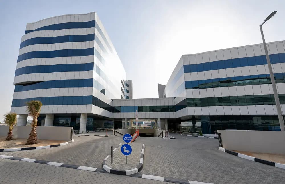 Modern office building exterior in Dubai with glass facades, entrance road, palm tree, and bright daytime sky.