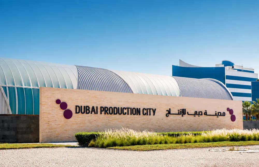 Dubai Production City entrance signage with English and Arabic text, landscaped greenery, and modern district buildings behind.
