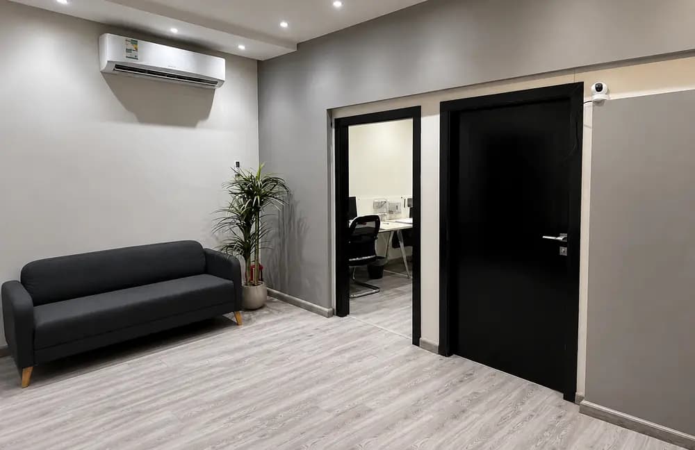 Riyadh office interior with grey sofa, plant, black doors, glass office opening, air conditioner, and light wood flooring.