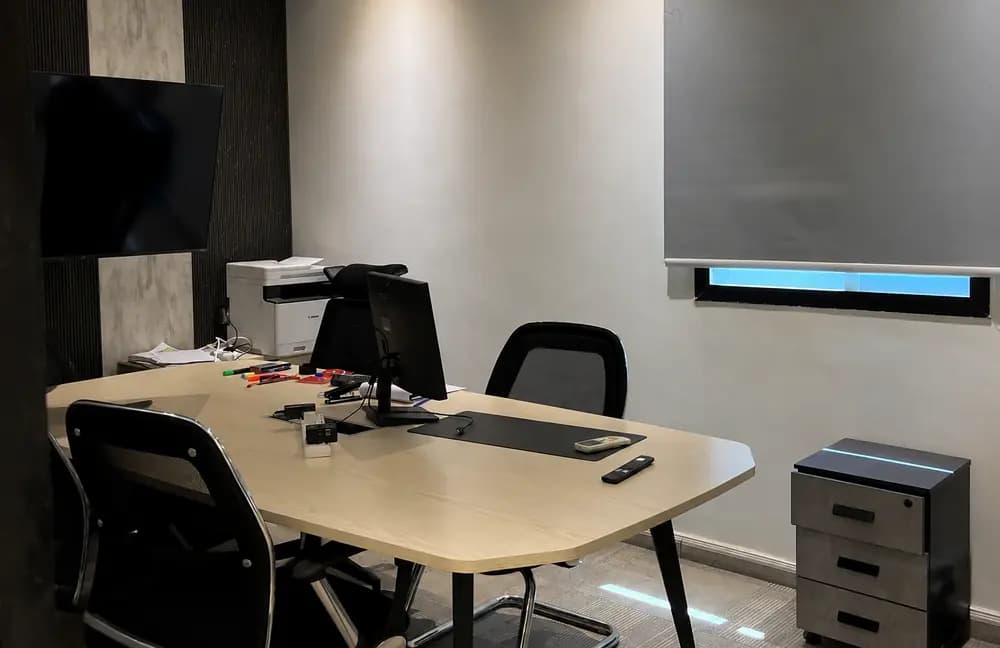 Riyadh office workroom with meeting desk, black chairs, computer monitor, printer, window blind, and grey interior walls.