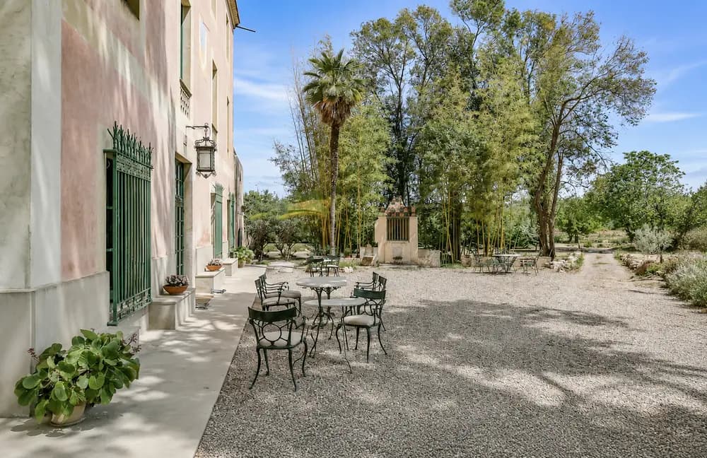 Courtyard beside the Cosido Farm farmhouse in Tarragona with outdoor tables, green window bars, gravel paths, trees, and garden shade.