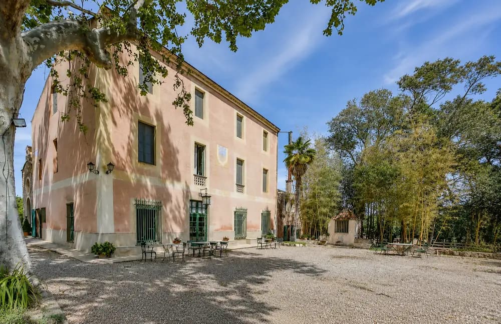 Pink historic farmhouse exterior at Cosido Farm in Tarragona with green doors, gravel courtyard, trees, and outdoor tables.