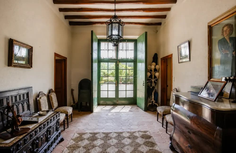 Historic entrance hall at Cosido Farm in Tarragona with green double doors, wooden beams, tiled floor, framed artwork, and antique furniture.