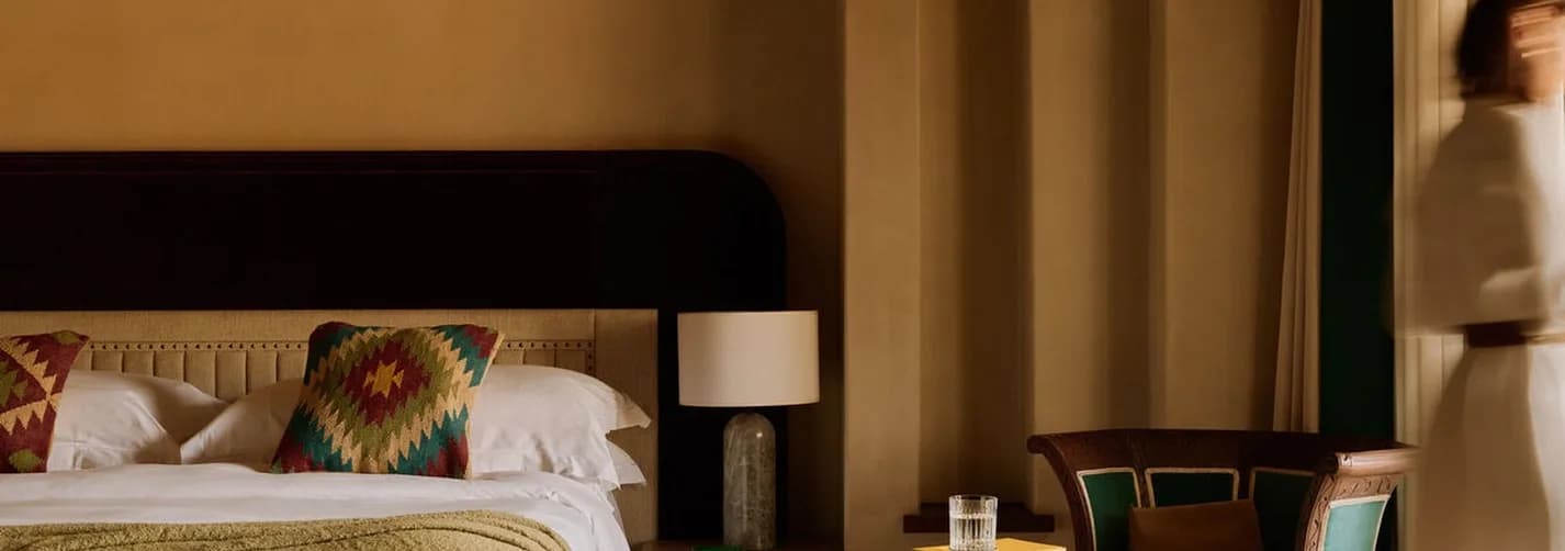 Luxury hotel bedroom in Dubai with warm interior styling, bed, patterned textiles, side table, handbag, and blurred guest movement.