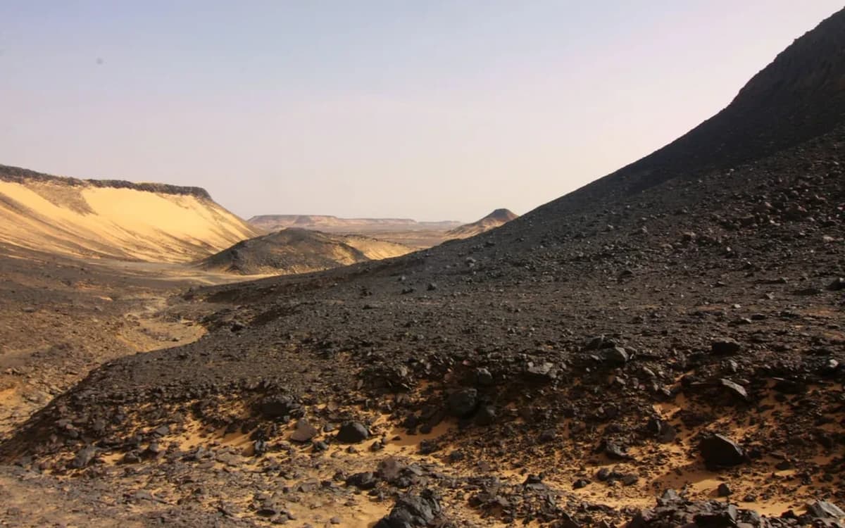 Black Desert landscape in Jordan with dark rocky slopes, sandy valley floor, and distant plateau light.