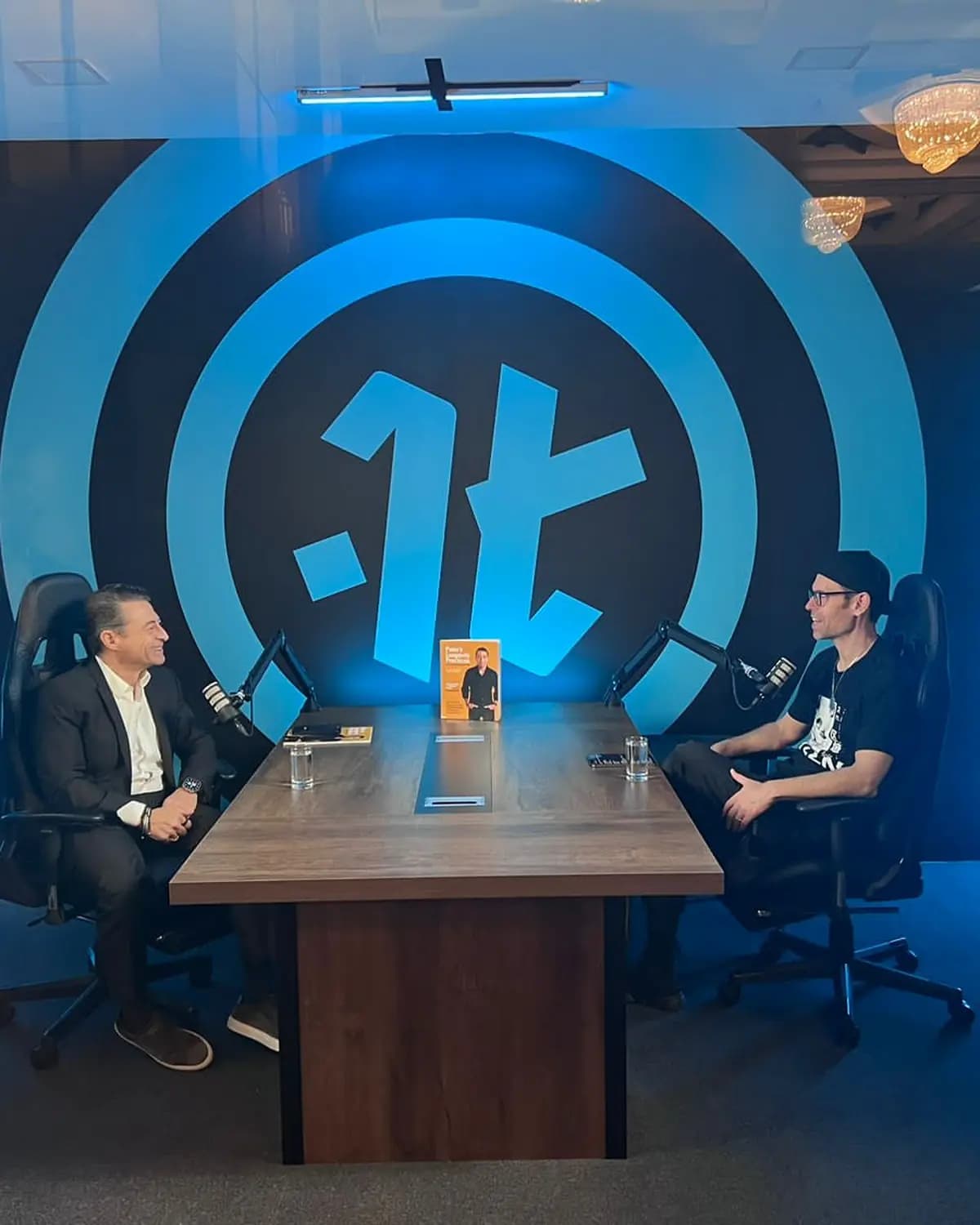 Impact Theory Podcast poster showing two speakers seated at microphones in a studio podcast setup.