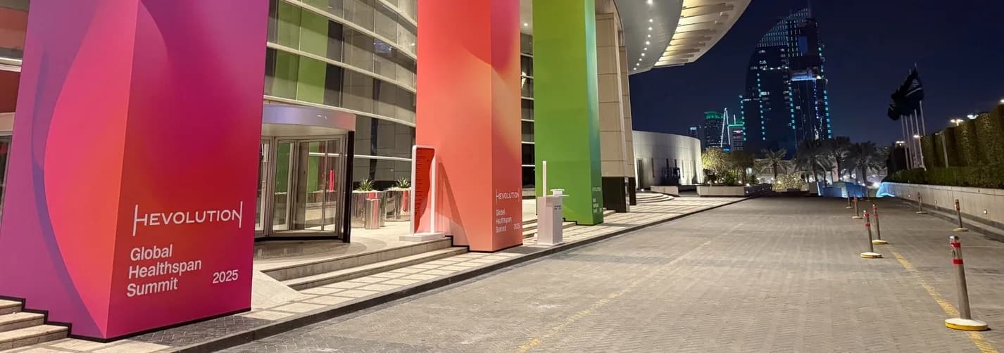 Night exterior of a Riyadh summit venue with colourful Hevolution Global Healthspan Summit 2025 entrance branding, city towers, and event arrival area.