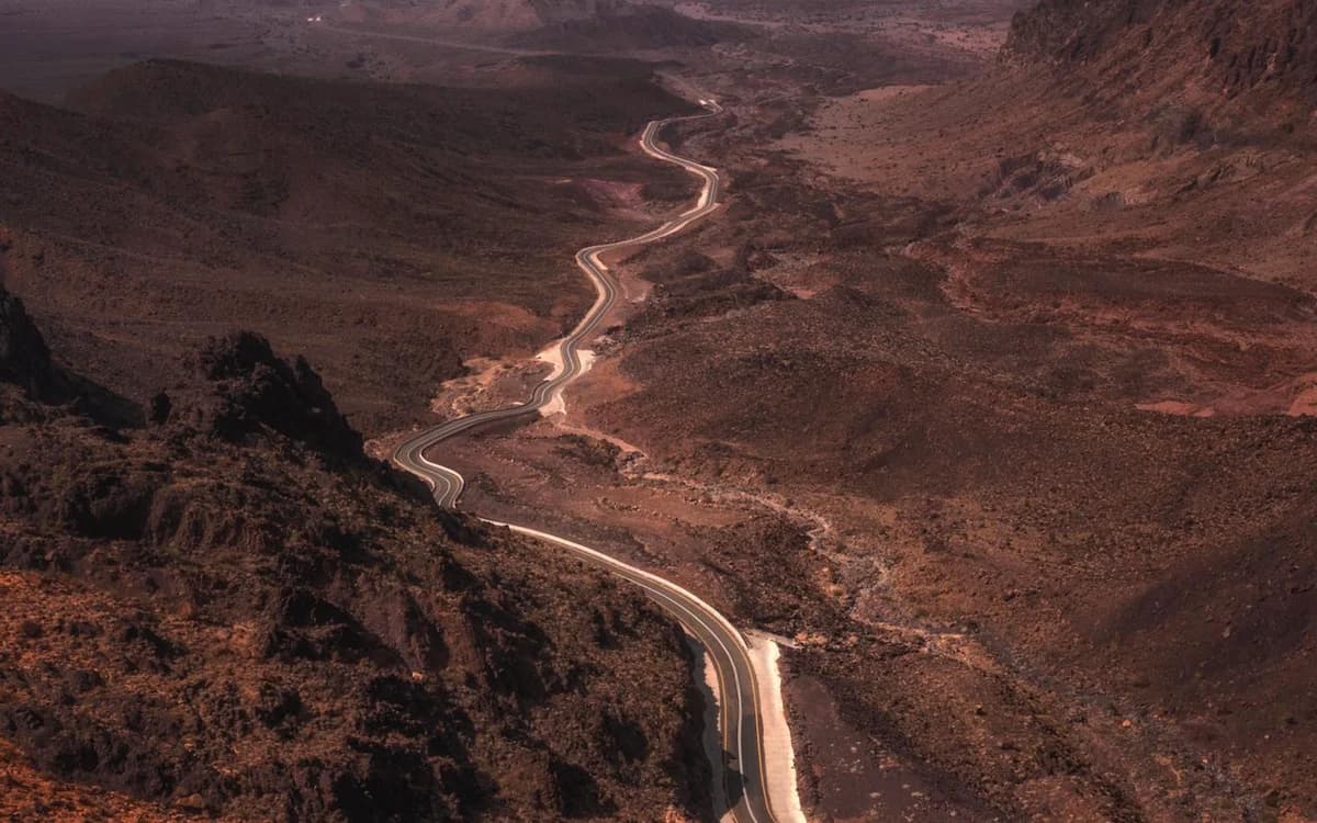 Winding desert road through AlUla’s rocky mountain landscape with dark volcanic terrain and canyon valleys.