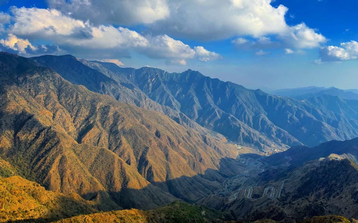 Aseer mountain range in Saudi Arabia with deep valleys, winding roads, rocky peaks, and dramatic cloud cover.