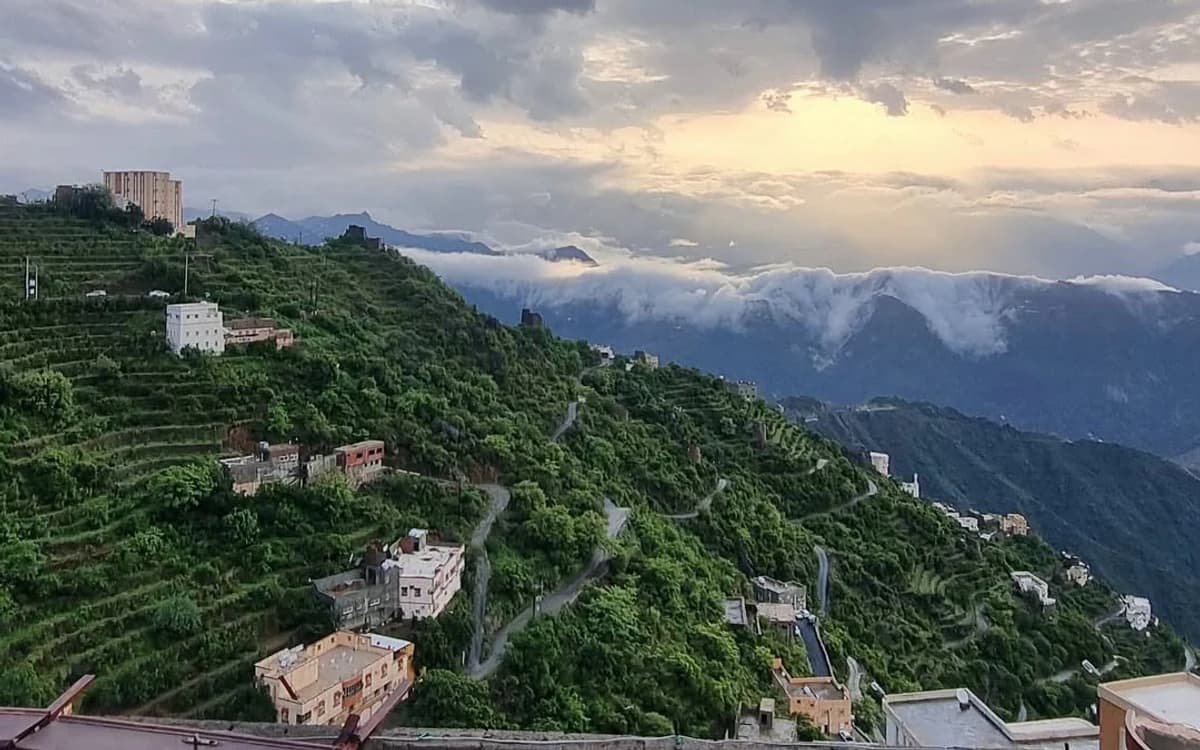 Green mountain terraces in Jazan with hillside roads, scattered buildings, clouds, and dramatic evening light.