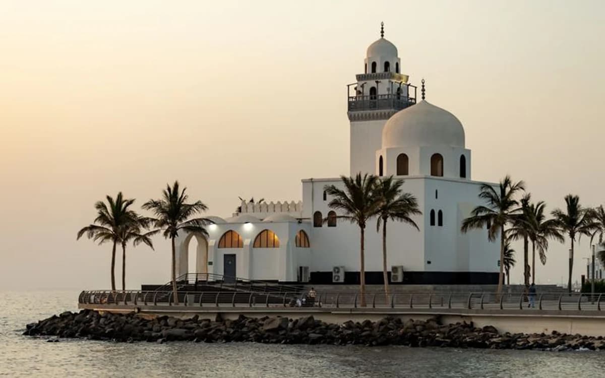 White waterfront mosque in Jeddah with palm trees, seawall, Red Sea water, and soft evening light.