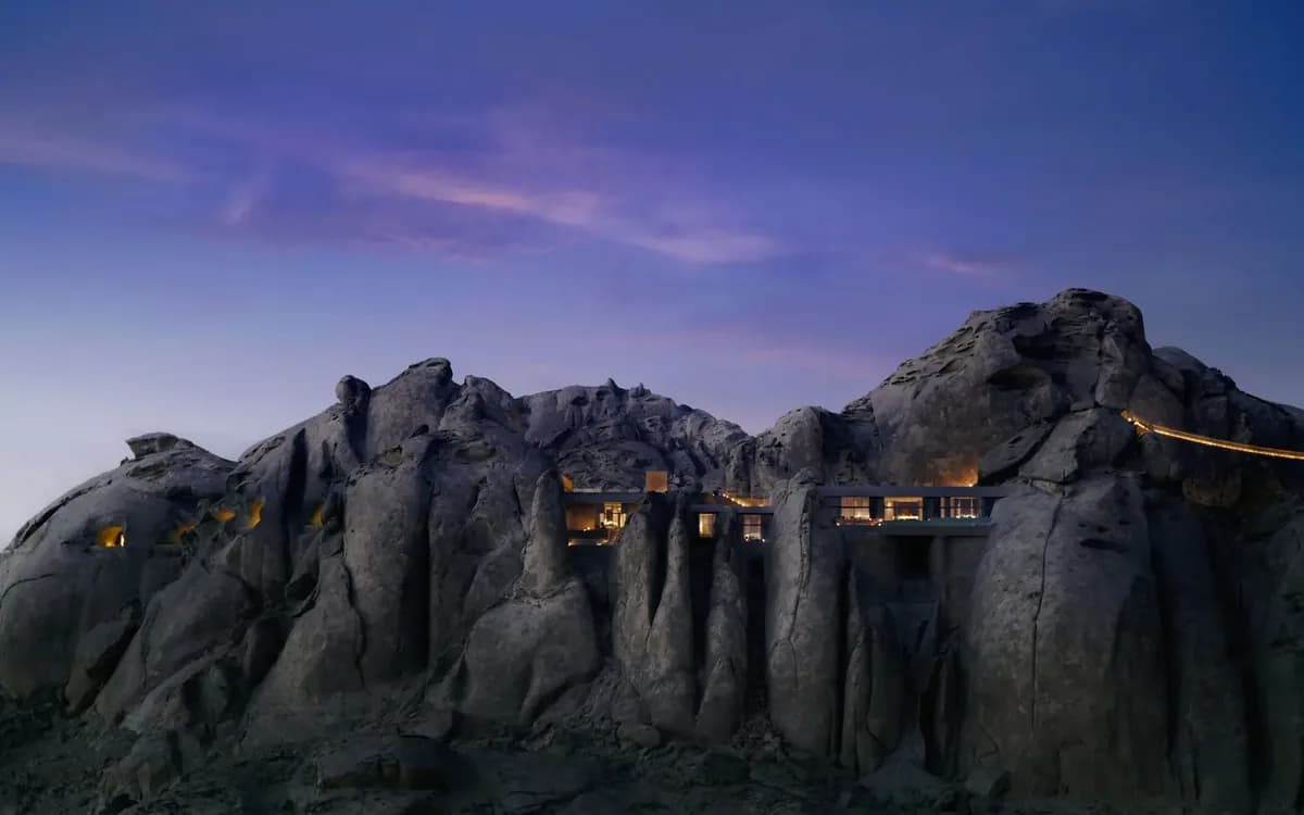 Wide exterior view of Desert Rock resort architecture integrated into sandstone boulders at dusk in Saudi Arabia.