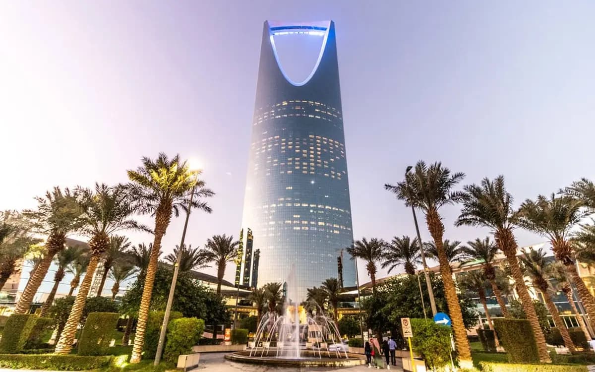 Kingdom Centre tower in Riyadh with palm trees, fountain, city lighting, and evening sky.