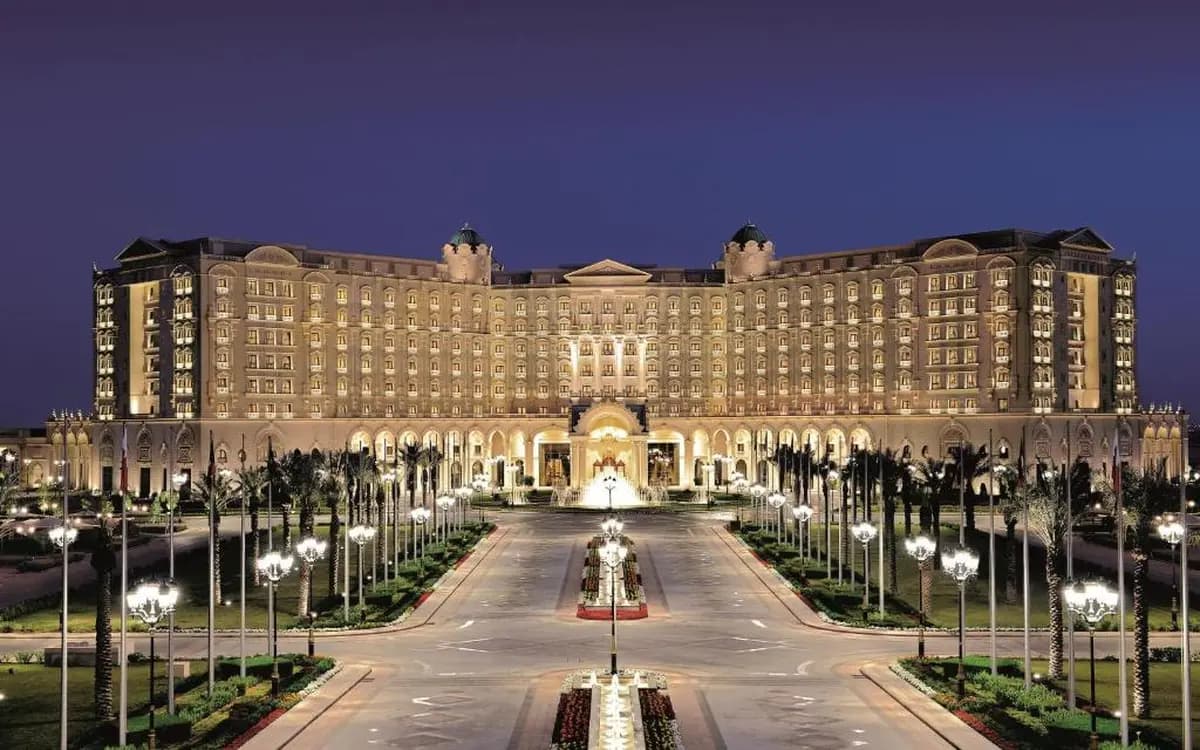Illuminated luxury hotel exterior in Riyadh with palm-lined entrance, fountain, and formal night setting.