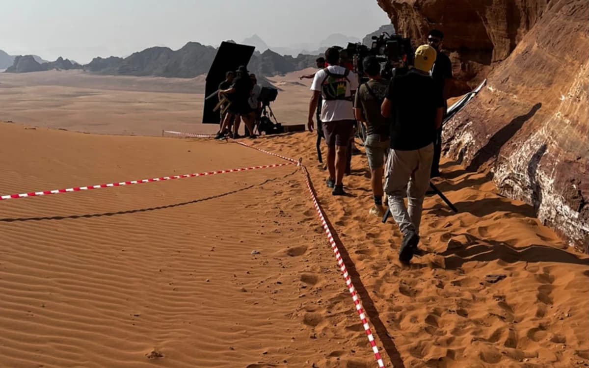 Film crew working on a desert location in Saudi Arabia with camera setup, black flag, marked access line, and sandstone cliffs.