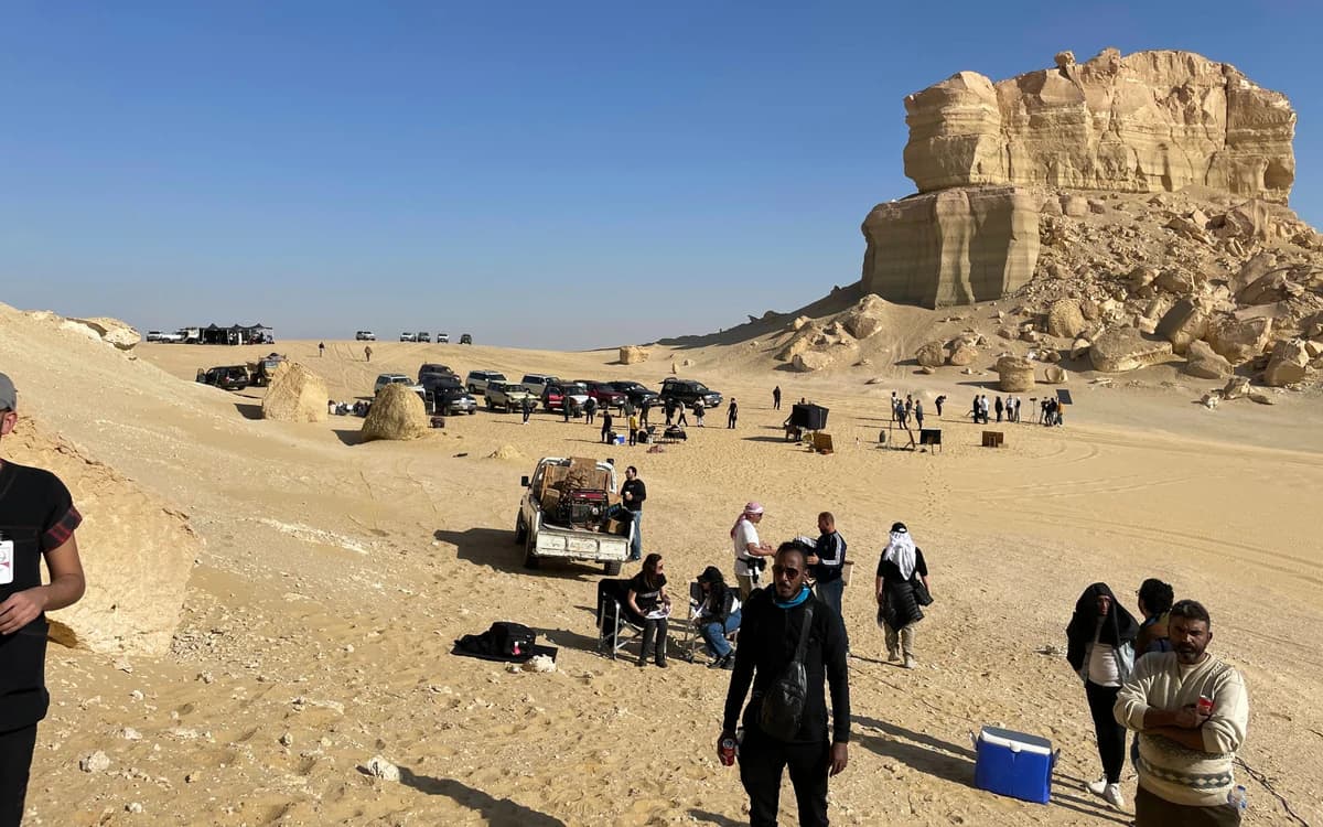 Desert film production setup in Saudi Arabia with crew, vehicles, equipment, and rock formations under clear sky.