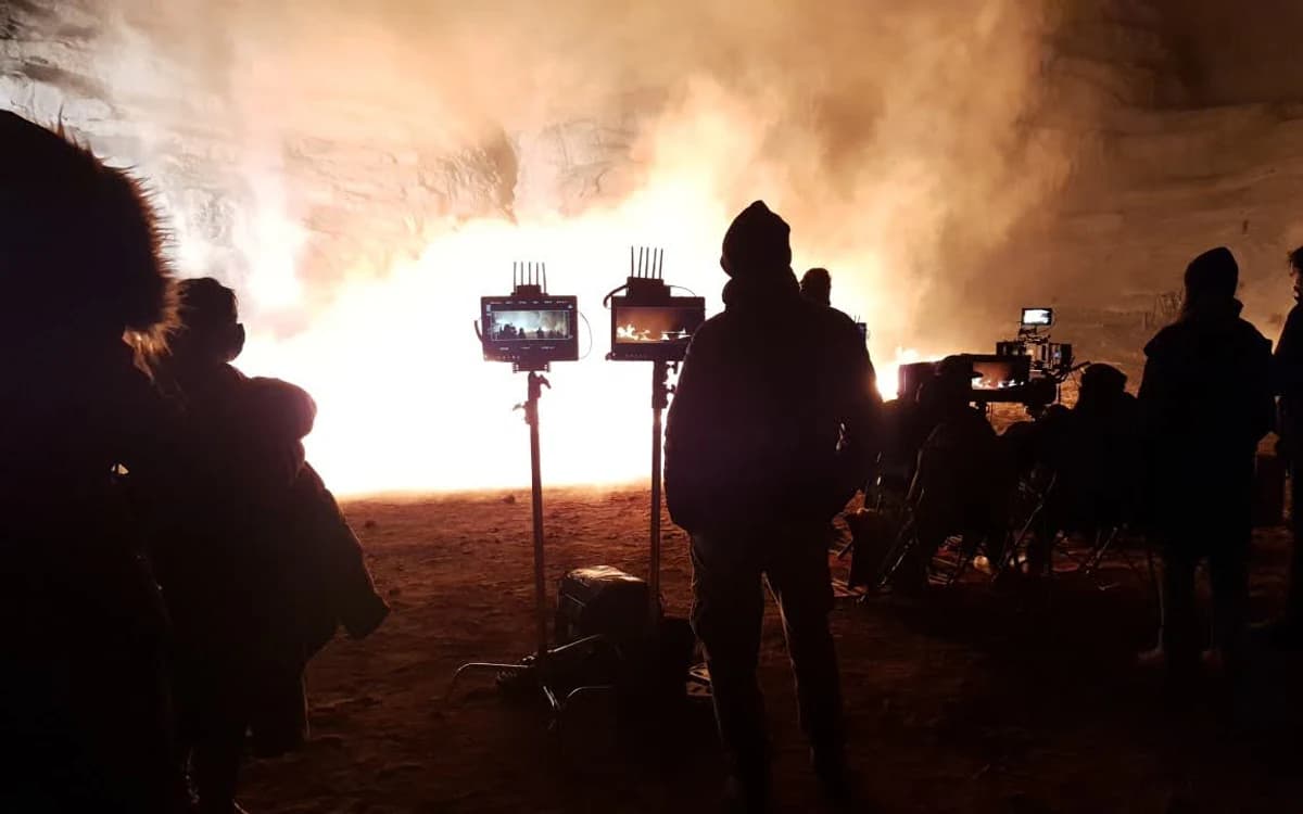 Night desert shoot in Saudi Arabia with silhouetted crew, camera setup, monitoring screens, smoke, and strong firelight.