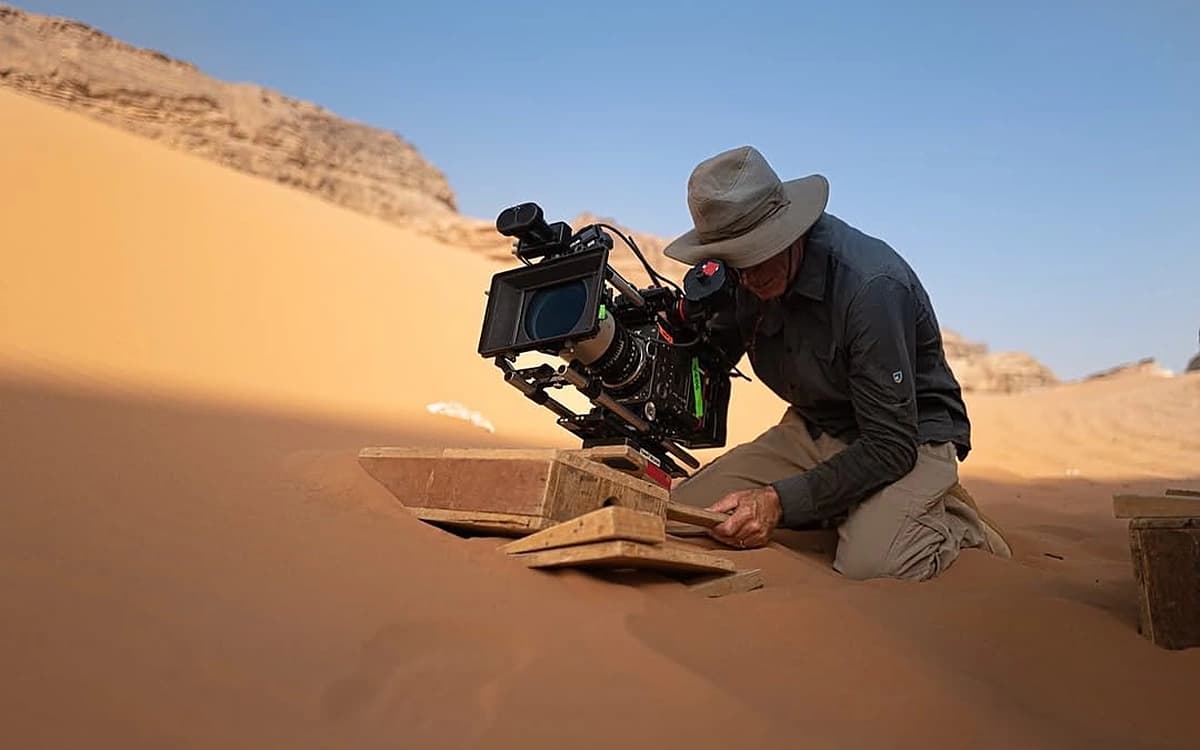 Camera operator setting up a cinema camera on sand in a Saudi desert with dunes and rocky landscape behind.
