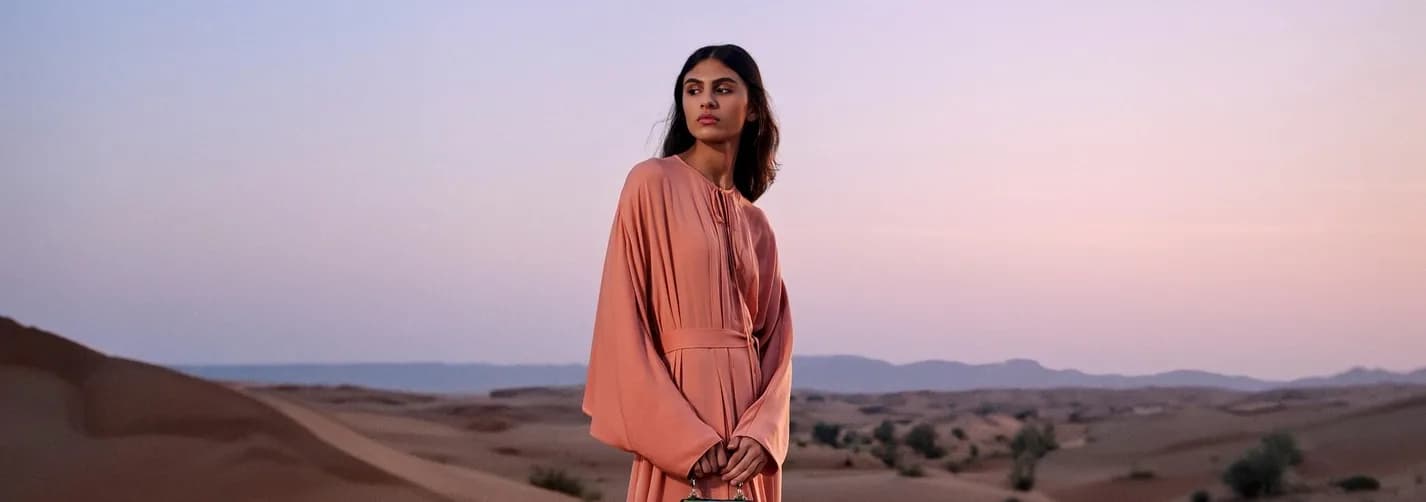 Fashion talent standing in the Sharjah Desert at dusk with sand dunes, distant rocky horizon, flowing dress, and handbag.