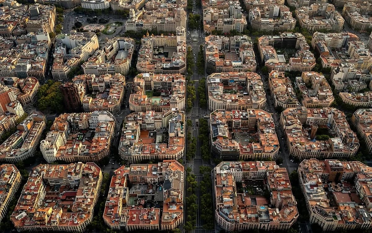 Aerial view of Barcelona's Eixample district with Cerdà blocks, tree-lined boulevard, rooftops, and dense city grid.