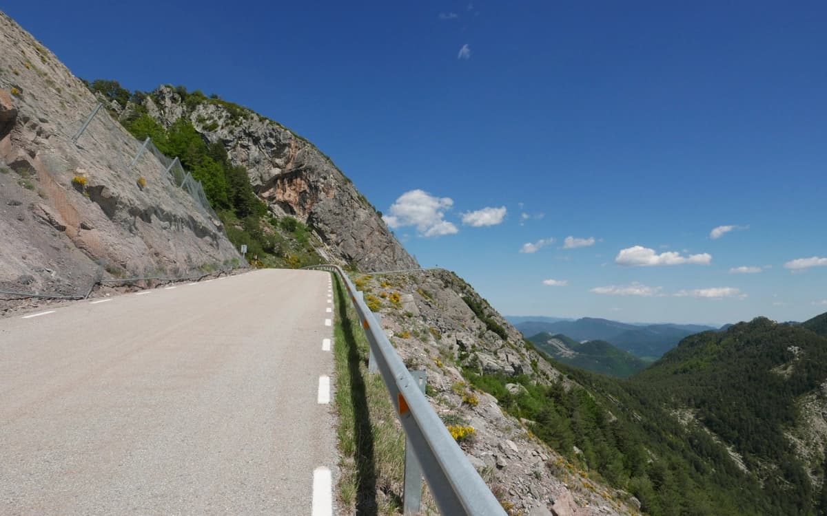 Cliffside mountain road in Catalonia with rocky slopes, guardrail, forested valley, and clear blue sky.