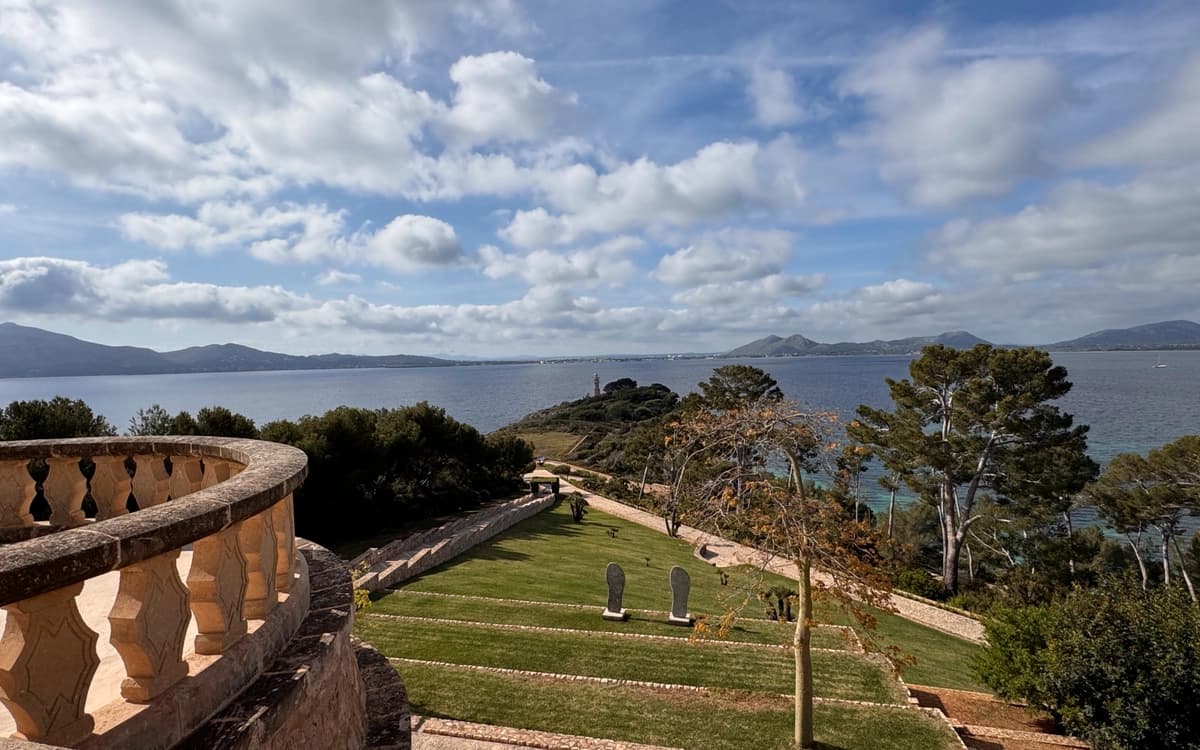 Historic estate terrace in Mallorca overlooking landscaped gardens, pine trees, coastline, and open Mediterranean water.