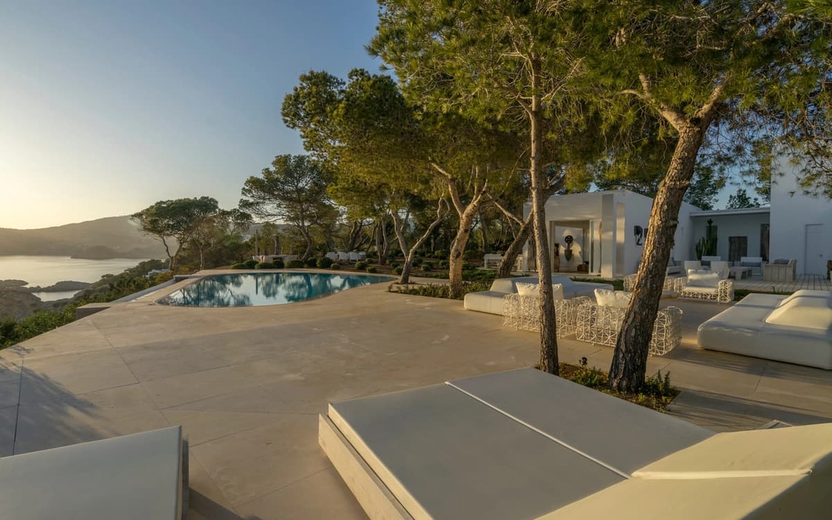 Cliffside villa terrace in Andratx, Mallorca with swimming pool, pine trees, sea view, white outdoor seating, and warm evening light.