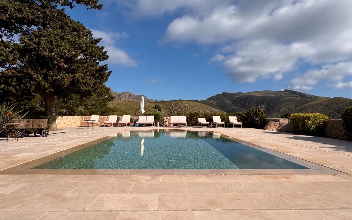 Luxury estate pool in Mallorca near Pollença with stone terrace, mountain backdrop, lounge chairs, trees, and open sky.