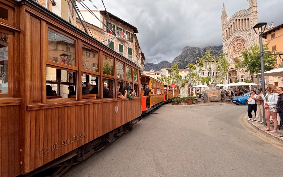 Historic wooden tram in Sóller, Mallorca passing through a town square with church facade, street cafés, pedestrians, and mountain backdrop.