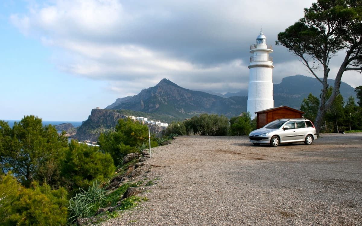 Cap Gros lighthouse near Sóller in Mallorca with cliffside parking area, pine trees, mountain backdrop, and Mediterranean coastline.
